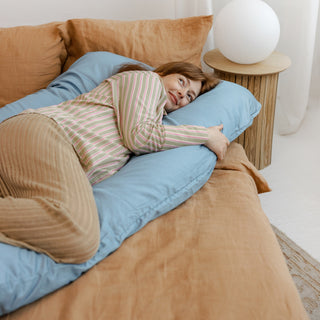 Pregnant woman relaxing on a bed, supported by a MINICAMP U-shaped comfort pillow with a natural sateen cotton cover in blue