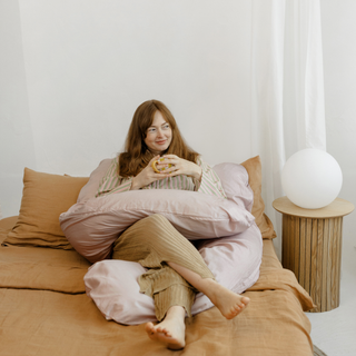 Moteris sėdi lovoje su puodeliu, apsupta kūno pagalvės, kuri suteikia komfortą ir atramą poilsiui